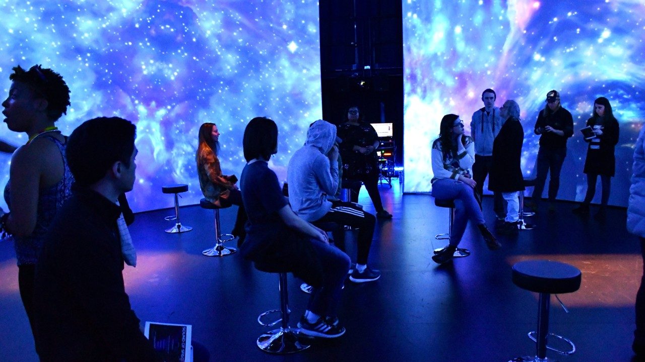 People listen to and experience "Sounds of Space" in the Cube at the Moss Arts Center. They're surrounded by a 360-degree swirling cosmic purple projection