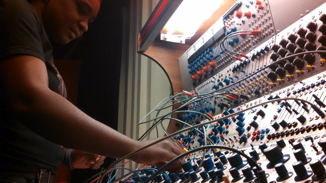 Afrofuturist artist Yvette Janine Jackson, a Black woman in a black T-shirt, plays a buchla