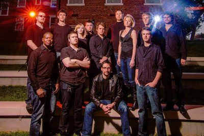 Thirteen musicians dressed in black pose outdoors on the amphitheatre steps behind Henderson Hall.