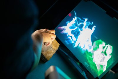 Exhibit-goer’s hand touches rock in front of a connected remote location projected on a screen. Photo by Rodney Kimbangu for Virginia Tech.