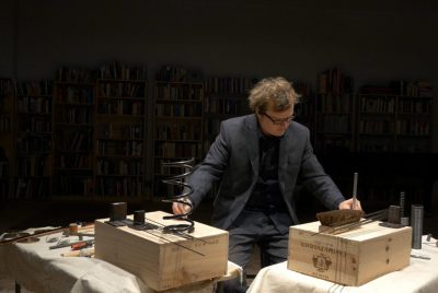 Ross Karre, a musician wearing a grey suit, stands behind two tables of various items such as springs, brushes, and rods on two wooden boxes.