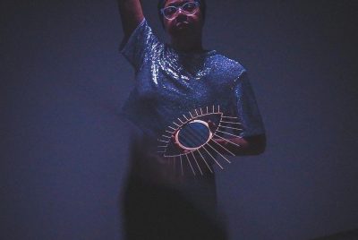 A woman standing with her arm stretched overhead, wearing a top wtih glitter, and large framed glasses, with a drawing of an eye and lashes over her other hand.