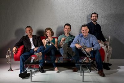 Five trumpet players, four men and one woman, sit on a red sofa holding their instruments.