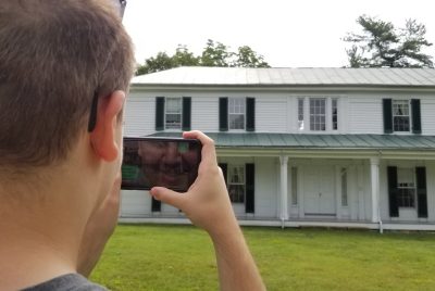 A student holds up a mobile phone and points it toward Solitude, Virginia Tech's oldest building - it's white with black shutters with a big front porch.