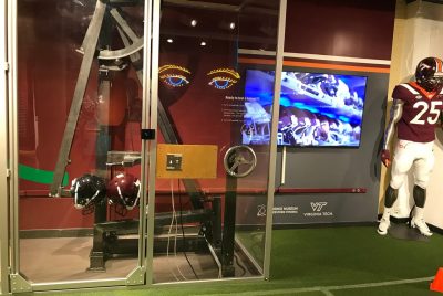 An exhibit at the Science Museum of Western Virginia includes a mannequin dressed in a Virginia Tech football uniform, a screen showing an image of many helmets, and a glass room where two helmets are pushed together using metal arms.